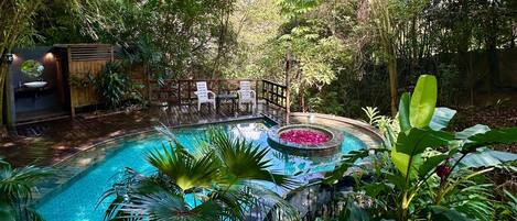 A swimming pool with a wooden deck, surrounded by lush greenery and a small waterfall feature.