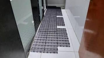 Um corredor estreito com piso de azulejos, porta de vidro à esquerda, área de pia com espelho e bancada, e porta à direita.