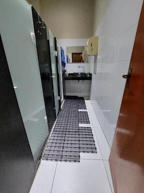 Um corredor estreito com piso de azulejos, porta de vidro à esquerda, área de pia com espelho e bancada, e porta à direita.