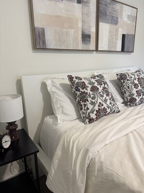 A neatly made bed with white bedding and floral patterned pillows, a bedside table with a lamp and an alarm clock, and abstract artwork on the wall. - 205 Bright Studio near Hospitals and Downtown (Indianapolis)