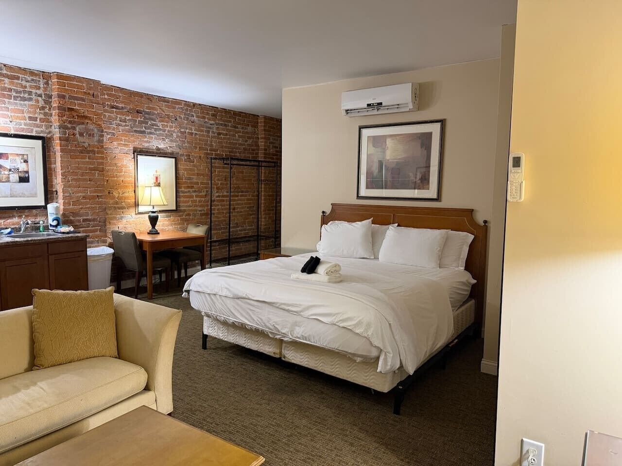 Une chambre d’hôtel avec un mur en briques, un lit avec une literie blanche, une tête de lit en bois, un bureau avec une chaise et une petite table.
