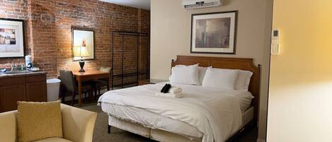 Habitación de hotel con pared de ladrillo, cama con ropa blanca, cabecera de madera, escritorio con silla y una mesita.