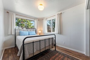 Quarto com cama, mesa de cabeceira, abajur e duas janelas com cortinas.