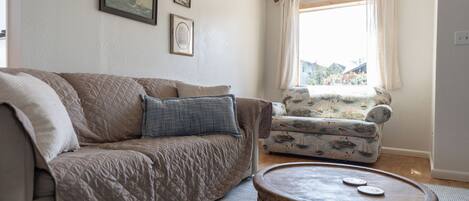 A living room with a beige sofa, a patterned armchair, a round coffee table, and a window with curtains.