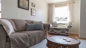 A living room with a beige sofa, a patterned armchair, a round coffee table, and a window with curtains.
