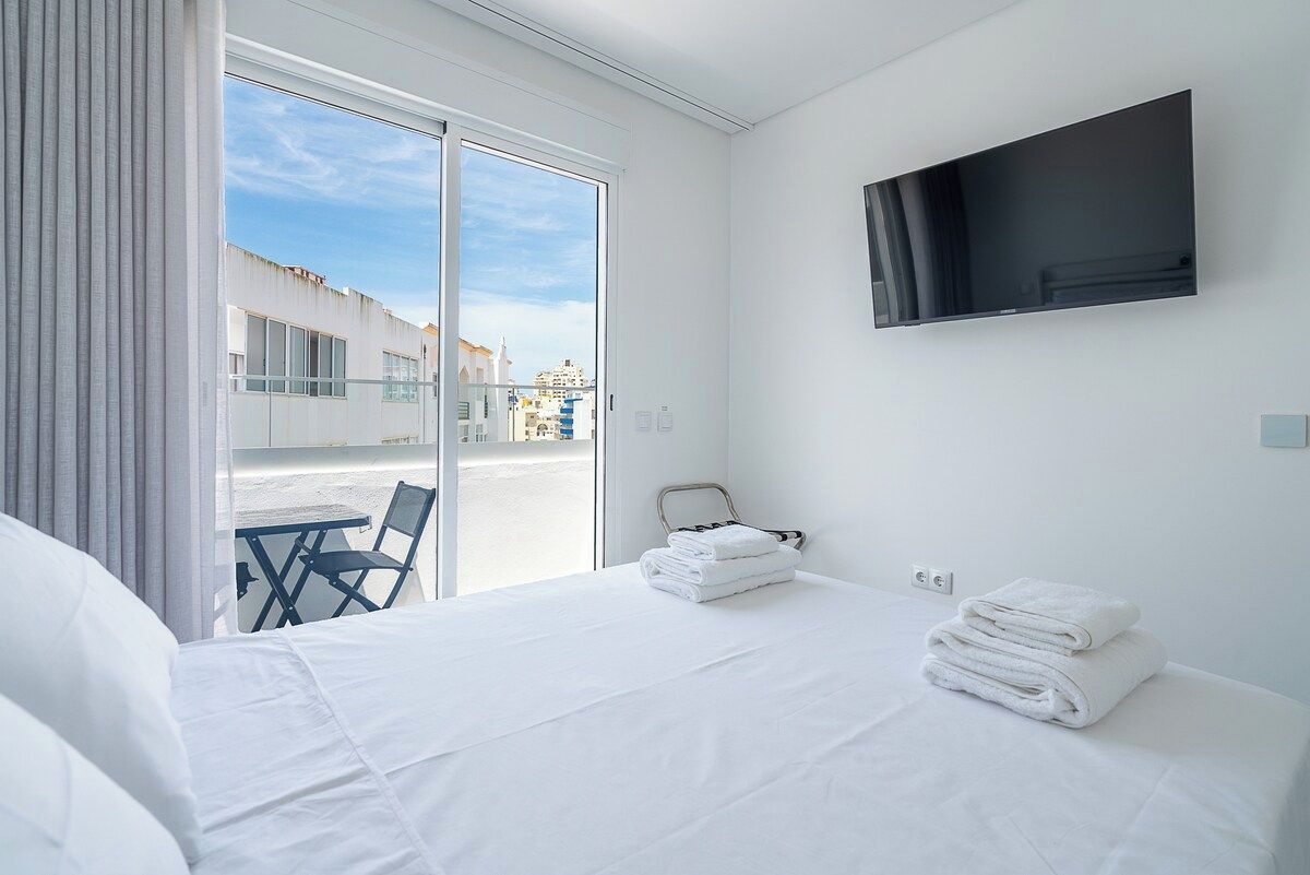 Ein Hotelzimmer mit Bett, Wandfernseher, Sessel und Tisch am Fenster.