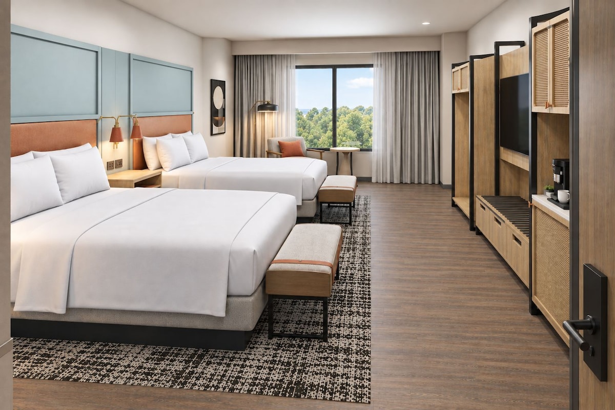 Habitación de hotel con dos camas, una mesita, una silla, televisión y un ventanal con vistas a árboles.