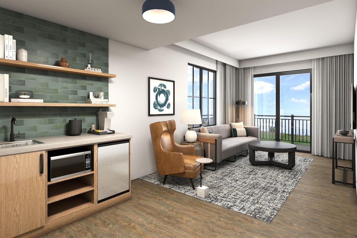 Una habitación de hotel moderna con cocina, zona de estar con sofá y sillones, mesa de comedor y balcón con vistas.