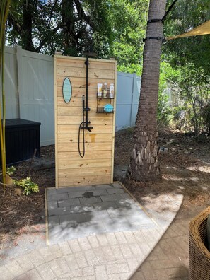 En udendørs bruseplads i træ med et brusehoved, et håndklædetørrestativ og en hylde med toiletartikler.