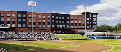 A baseball field with a modern stadium in the background, seating for spectators, and a clear sky.