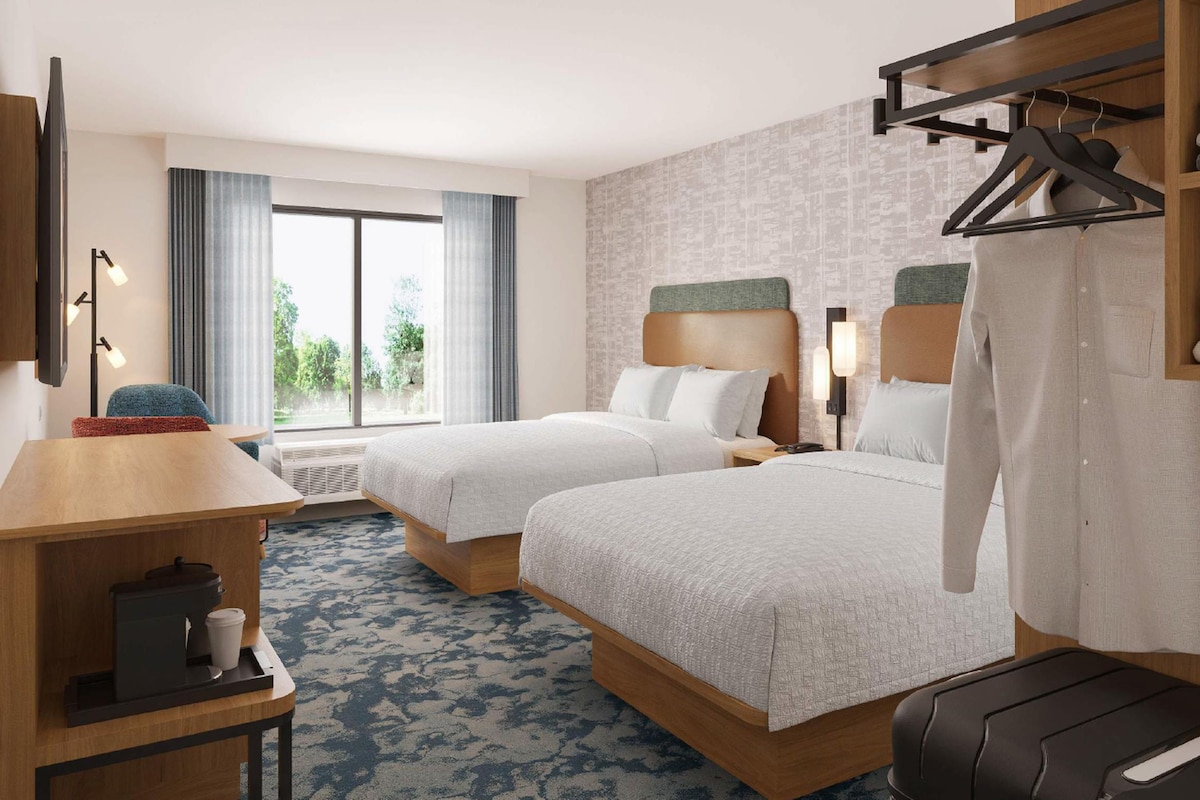 Quarto de hotel com duas camas, uma escrivaninha, uma máquina de café, uma mala e vista para o exterior.