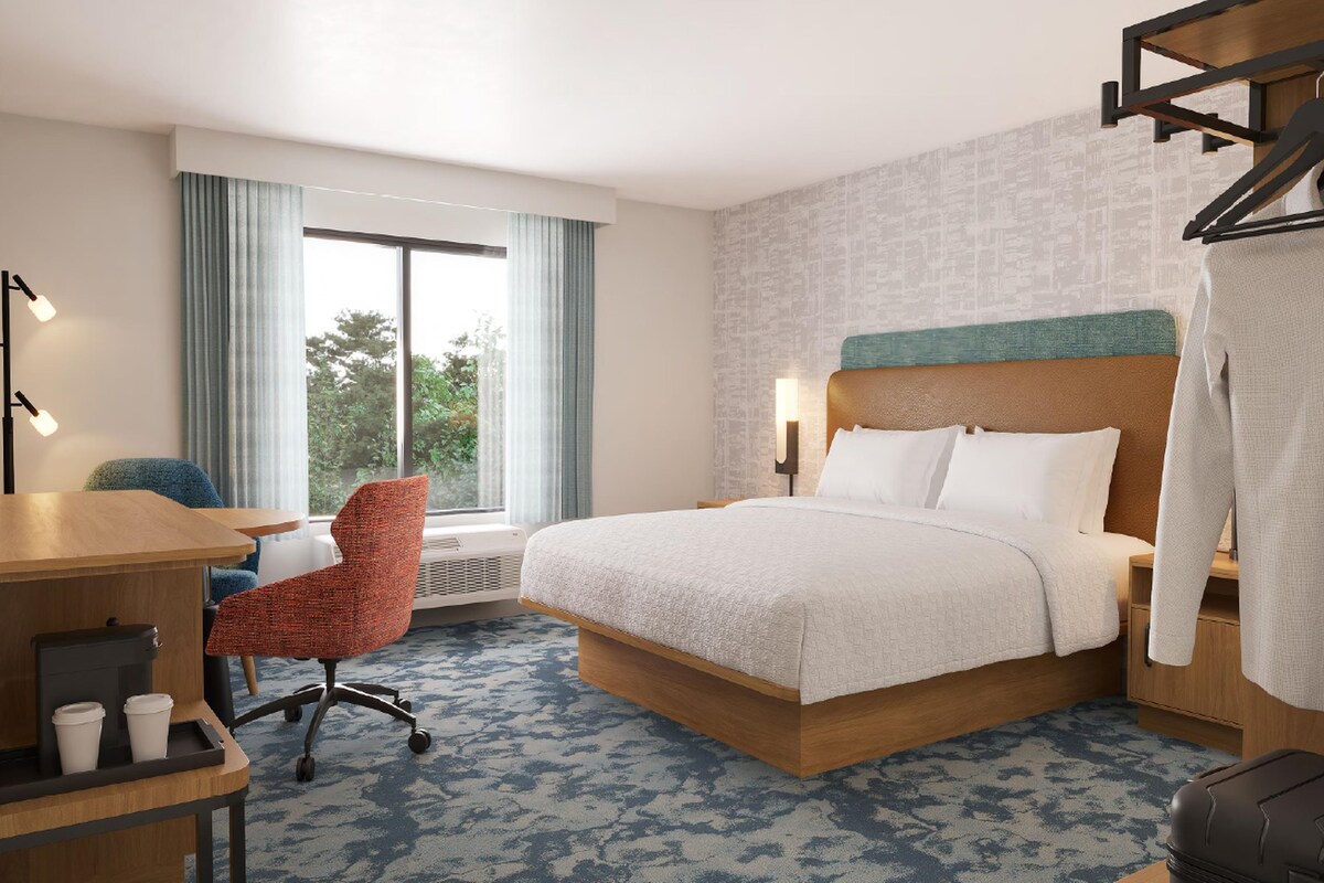 Quarto de hotel com cama, escrivaninha com cafeteira, cadeira, mala e janela com cortinas.