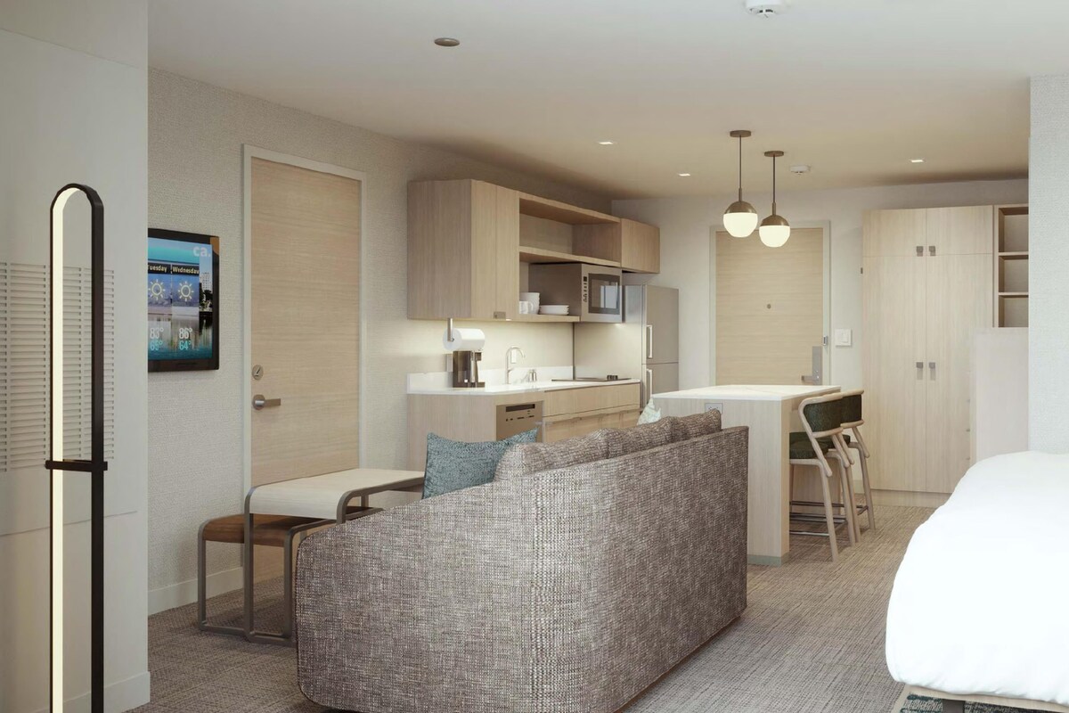 Quarto de hotel moderno com cozinha compacta, sofá, área de refeições e cama.