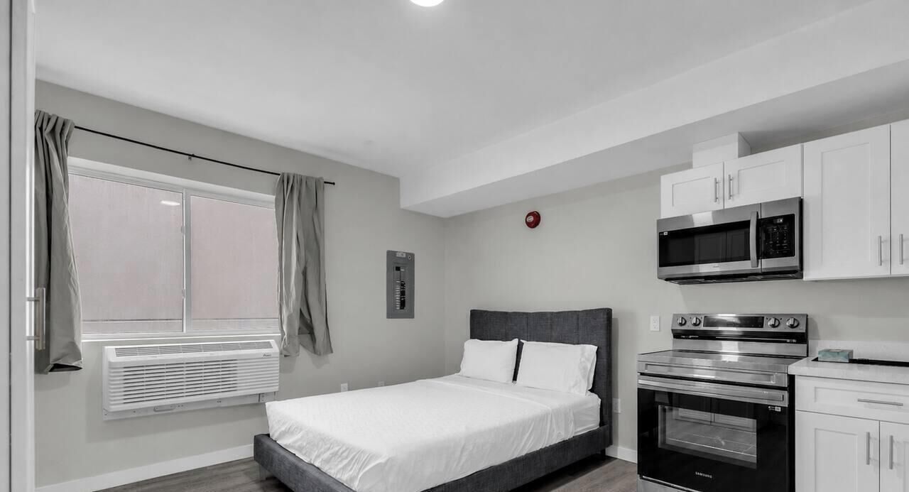 Ein Schlafzimmer mit einem Bett, einem Fenster mit Vorhängen, einer Wandheizung, einer Mikrowelle und einem Backofen.
