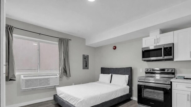 A bedroom with a bed, a window with curtains, a wall-mounted heater, a microwave, and an oven. - The Vista Suite • Studio • DT Oshawa (Oshawa)