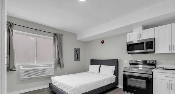 A bedroom with a bed, a window with curtains, a wall-mounted heater, a microwave, and an oven. - The Vista Suite • Studio • DT Oshawa (Oshawa)