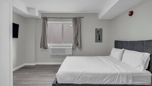 A hotel room with a large bed, a window with curtains, a wall-mounted air conditioning unit, and a fire alarm. - The Vista Suite • Studio • DT Oshawa (Oshawa)