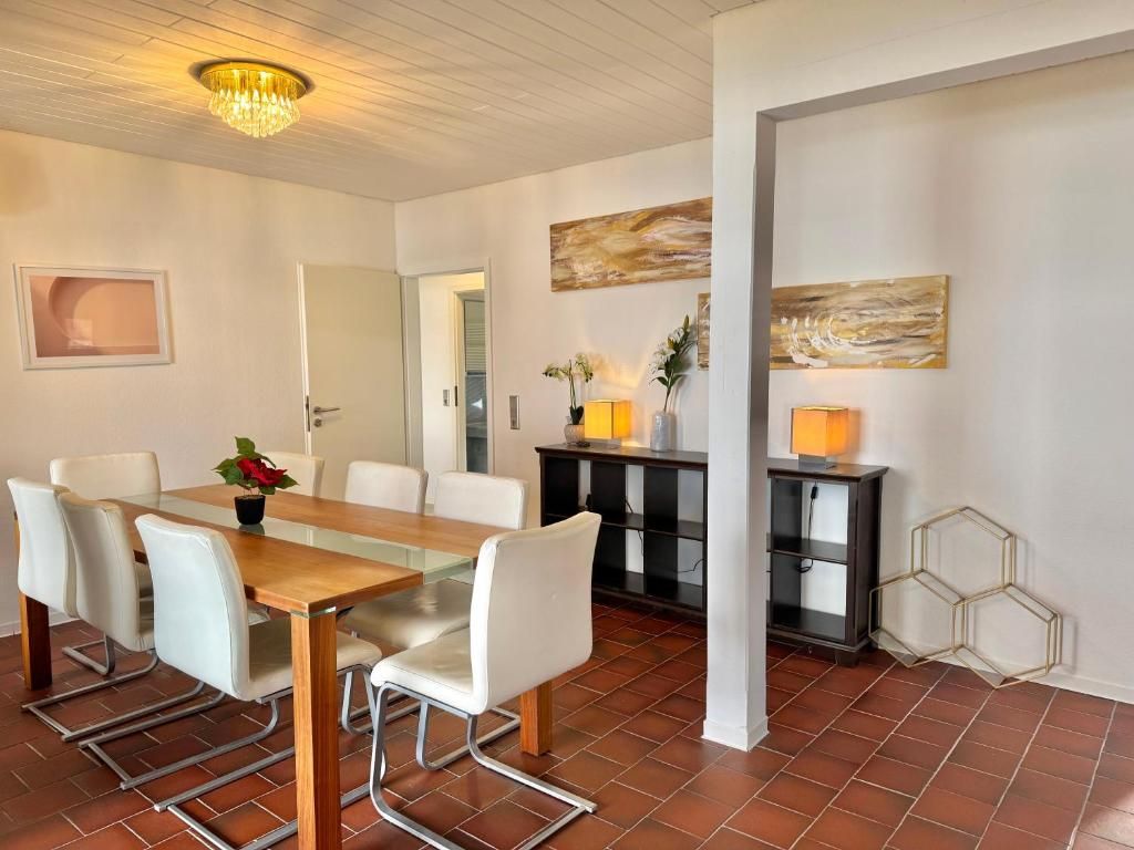 Un comedor con mesa de madera, sillas blancas y una araña. Se observa un aparador de diseño hexagonal y un cuadro en la pared.