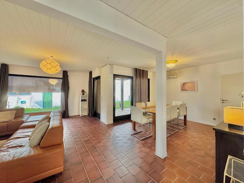 A spacious living area with a sofa set, dining table, and chairs, a ceiling with a unique light fixture, and a view through a glass door.