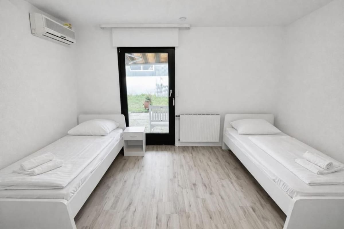 A small, clean room with two single beds, a small table, and a window with a view of the outside.