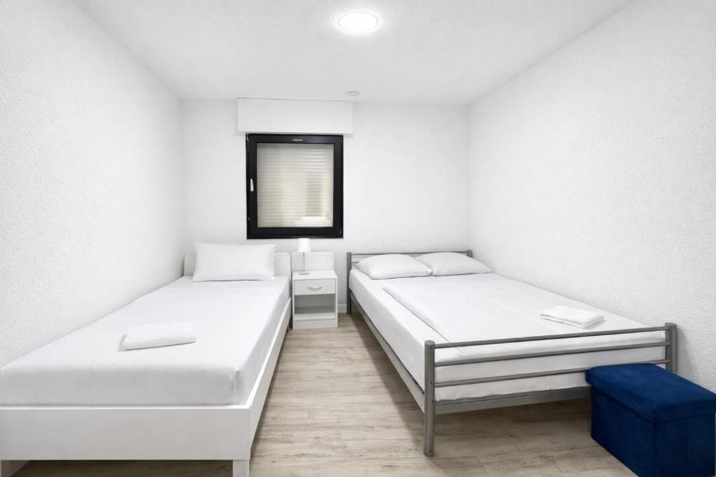 Una habitación pequeña y limpia con dos camas individuales, una mesita de noche, una ventana con persianas y un banco azul.