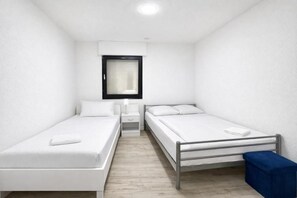 A small, clean room with two single beds, a nightstand, a window with blinds, and a blue bench.