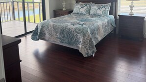 Quarto com cama, ventilador de teto, mesa de cabeceira com luminária, um quadro e vista para a água através de uma porta de correr.