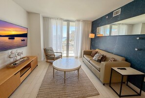 A living room with a sofa, a chair, a coffee table, and a TV displaying a sunset view. - Luxury Beach Resort 1BR Sunset View & Beach access (Hallandale Beach)