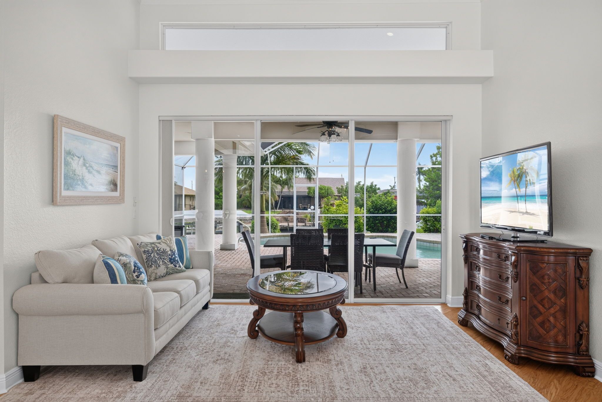 A living room with a sofa, coffee table, and TV. There is a sliding glass door leading to an outdoor patio with a pool and chairs.