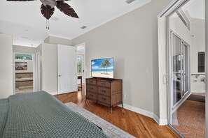 A bedroom with a bed, a dresser, a television, and a sliding glass door leading to a balcony. - Heated Pool home with Amazing Gulf Access in Southeast Cape! (Cape Coral)