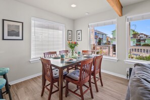 A dining area with a glass table, wooden chairs, and a view of neighboring houses through large windows. - Sunkissed 3BR - Oceanview Beach Home (Pacific Beach)