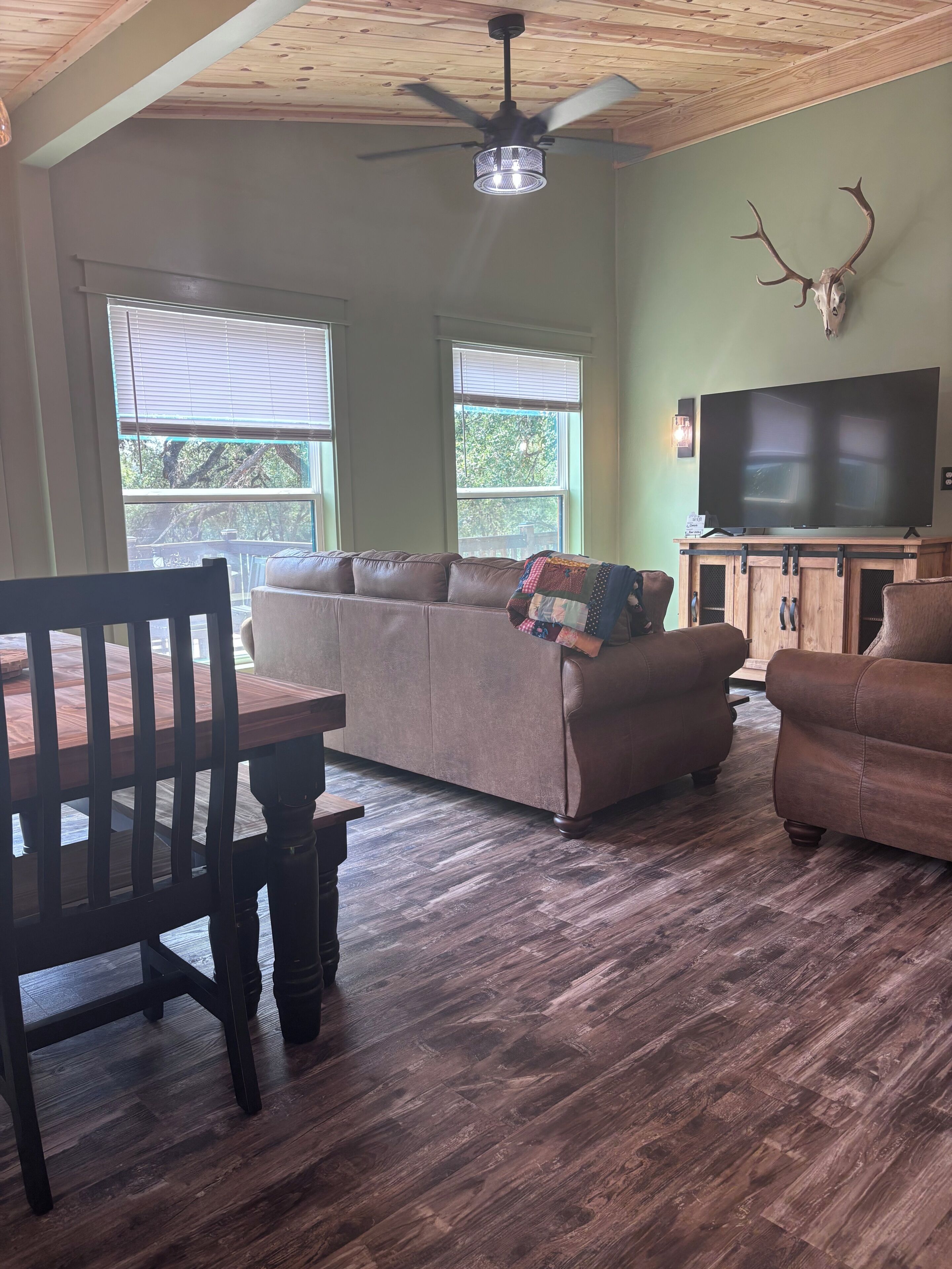 A living room with a wooden ceiling, a ceiling fan, a television, a sofa, a coffee table, and a dining table with chairs.