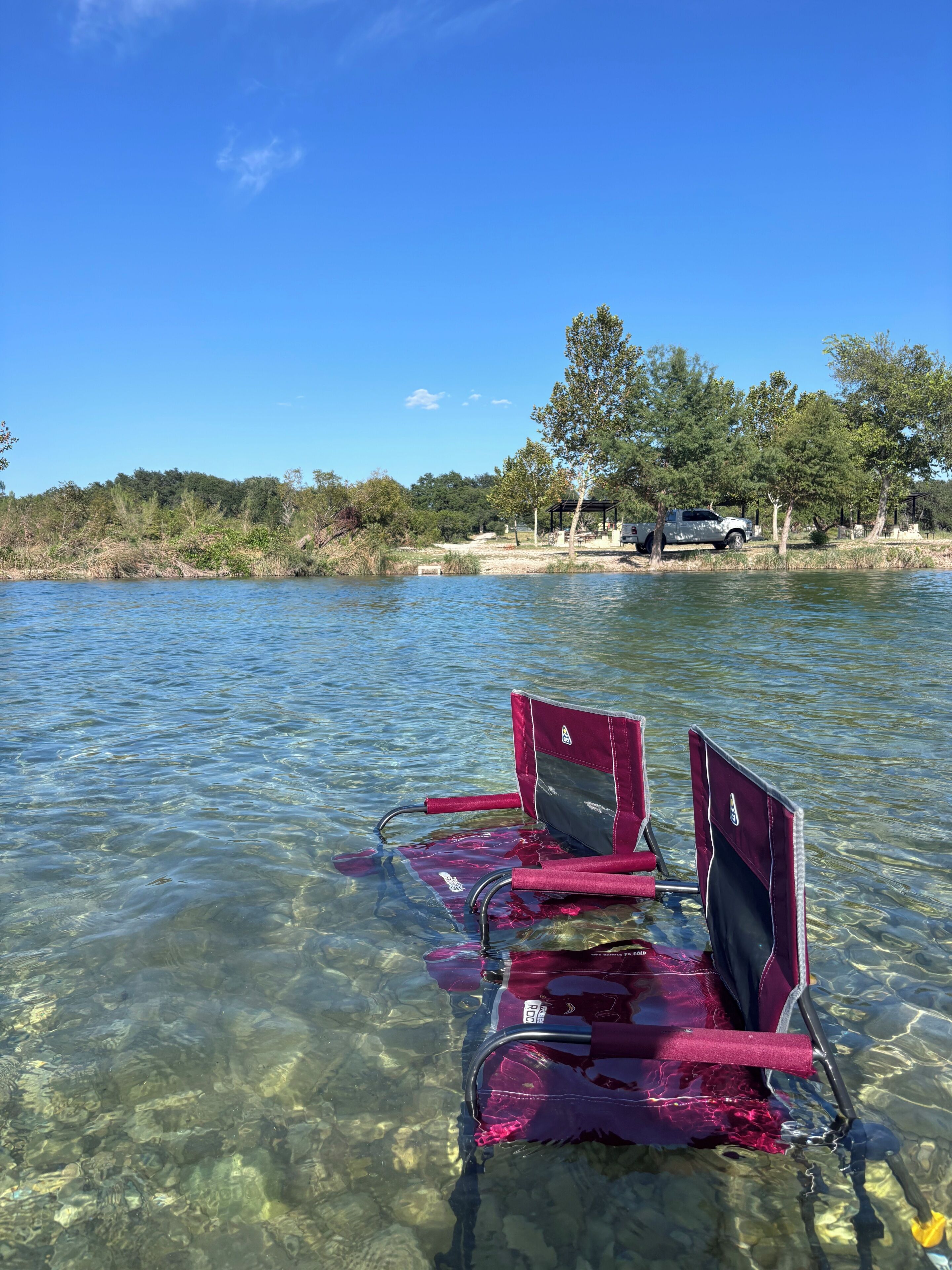 Un lac aux eaux limpides, avec deux chaises rouges flottant à sa surface, entouré d’arbres et avec un véhicule visible au loin.