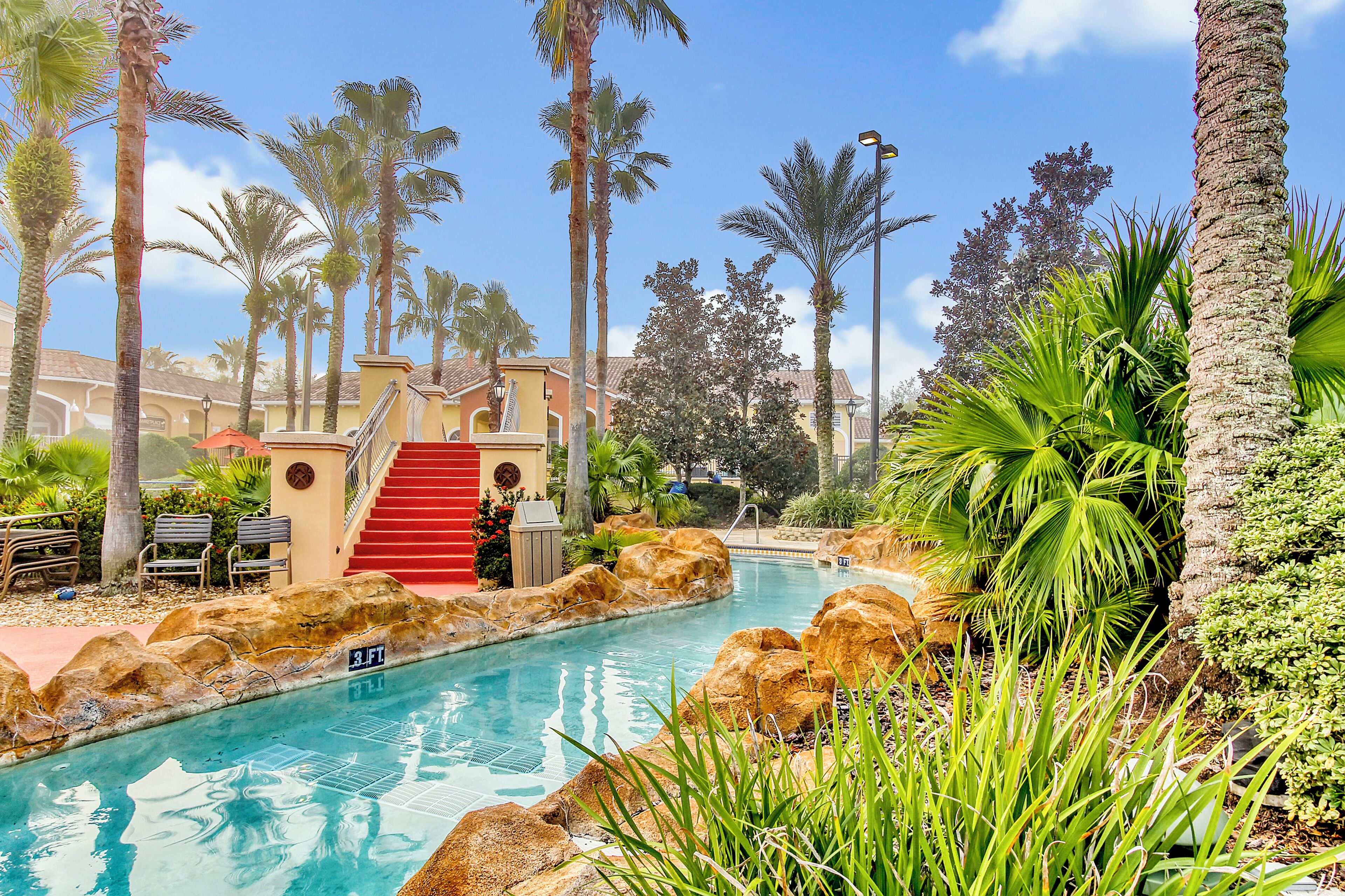 A pool area with a red staircase leading to a building, surrounded by palm trees and rocks.