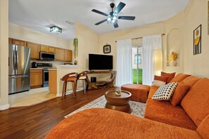 Ein modernes Wohnzimmer mit einer Couch, einem Couchtisch und einem Fernseher. Es gibt eine Küchenzeile mit einem Kühlschrank und einer Mikrowelle.