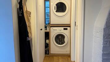 A laundry room with two stacked washing machines, a hanging rack with clothes, and a wooden floor.
