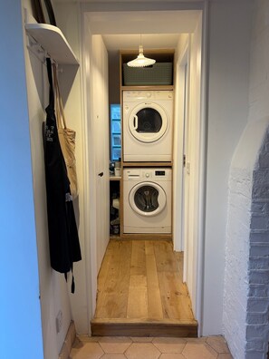 A laundry room with two stacked washing machines, a hanging rack with clothes, and a wooden floor.