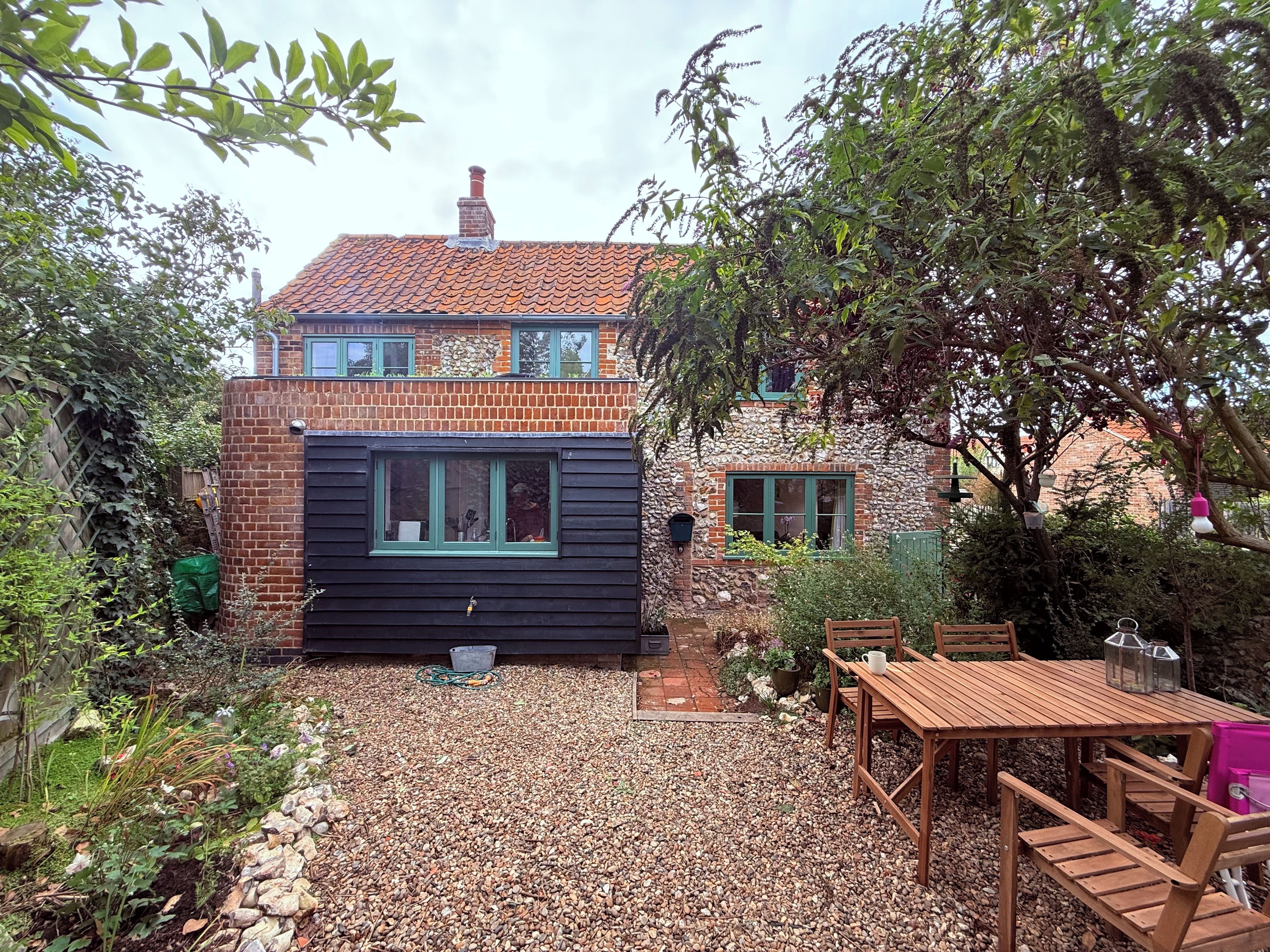 A two-story house with a brick facade, a small attached wooden structure, and a gravel patio with wooden furniture.