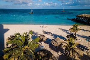 A beach with clear turquoise waters, a couple walking on the sand, and thatched umbrellas. - AL318 -Resort Pool-Beach Tennis court- 2BR+ condo-Close to Palm Beach (Noord)