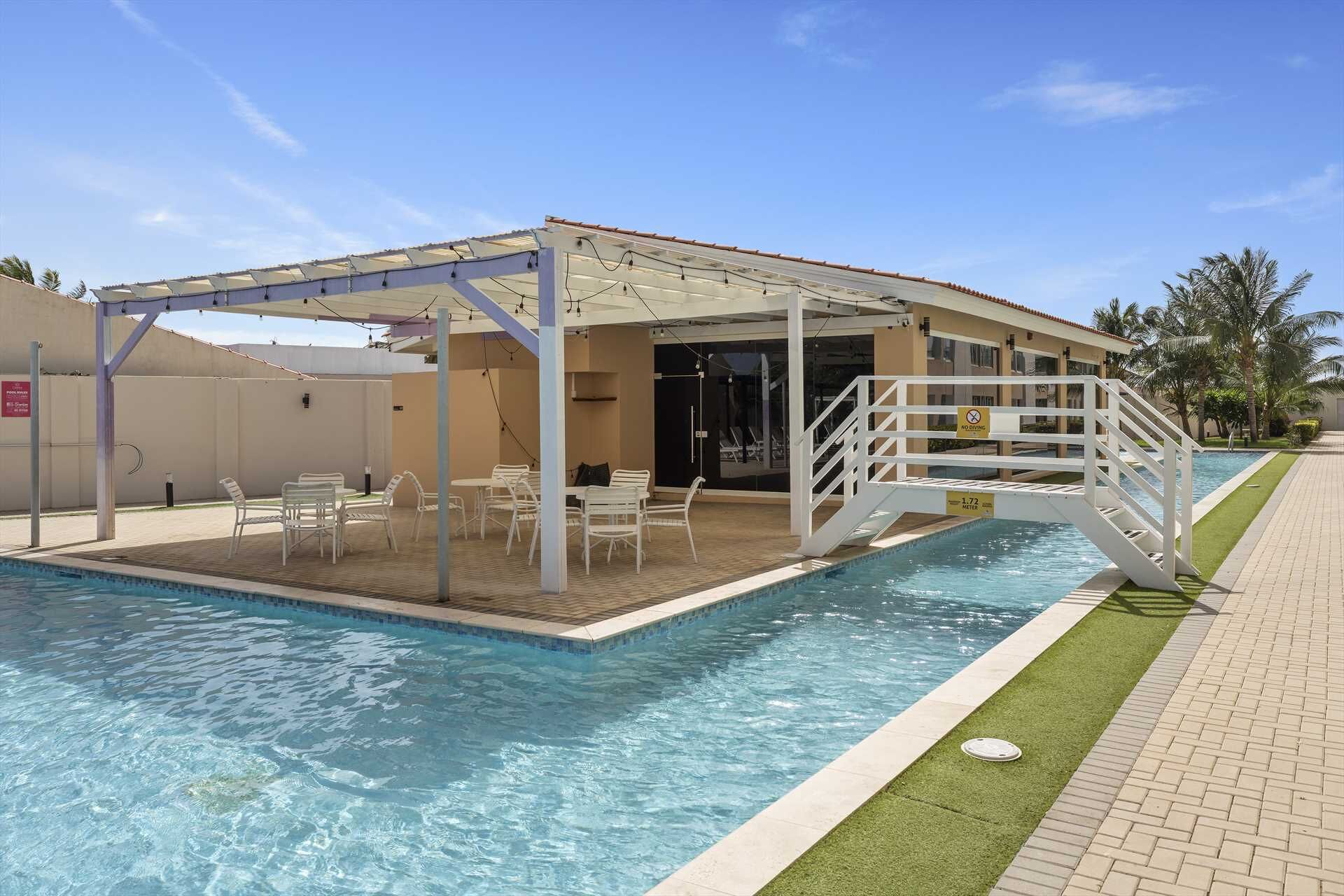 Ein Schwimmbecken mit überdachter Sitzgelegenheit, Liegen am Pool und einer Treppe, die zu einem Gebäude führt.