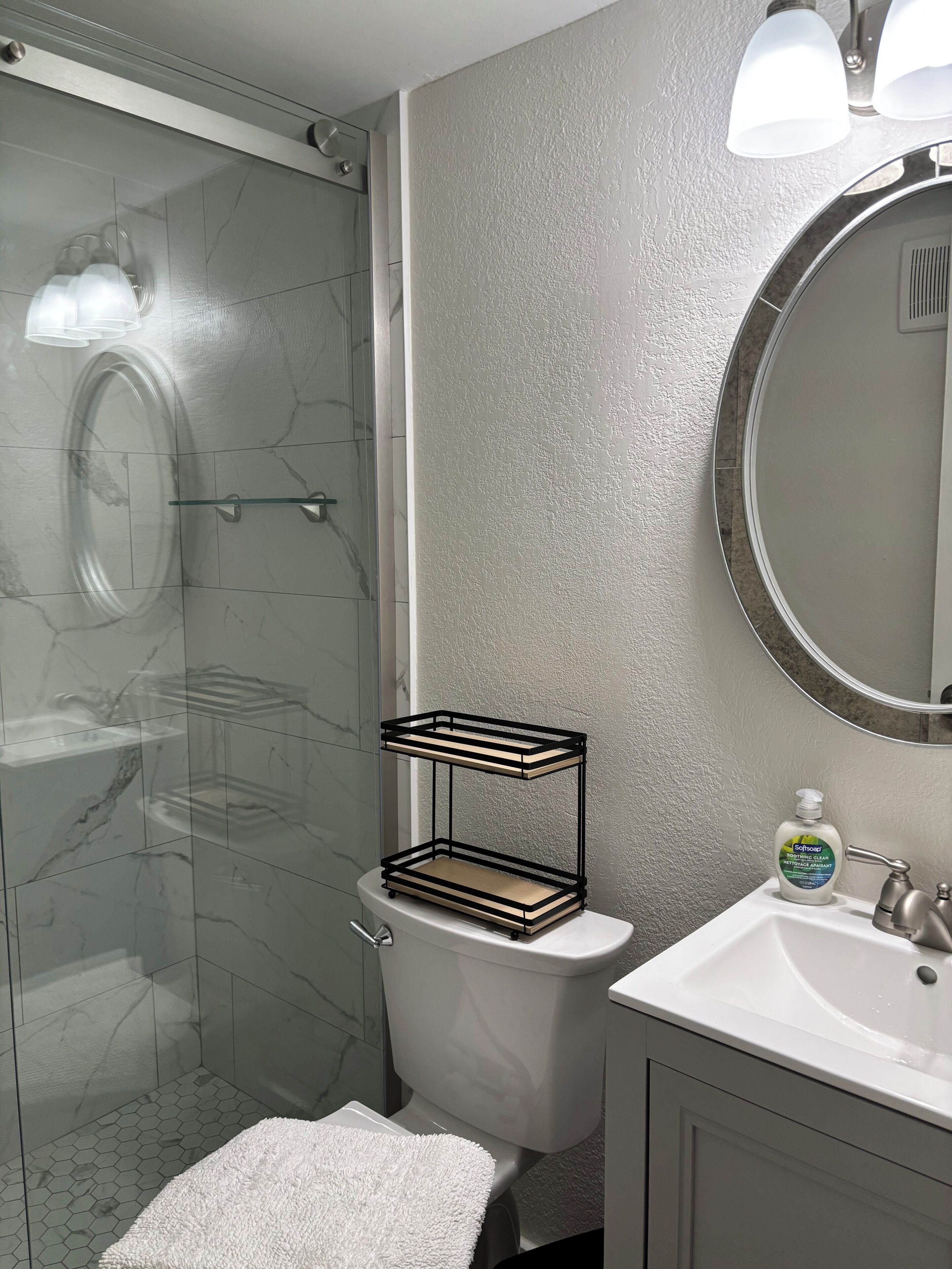 Ein Badezimmer mit Duschkabine aus Glas, Toilette, Waschbecken mit Seifenspender und einem Handtuch auf dem Boden.