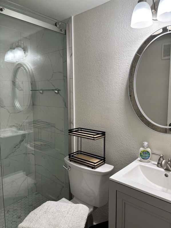 A bathroom with a glass shower enclosure, a toilet, a sink with a soap dispenser, and a towel on the floor.