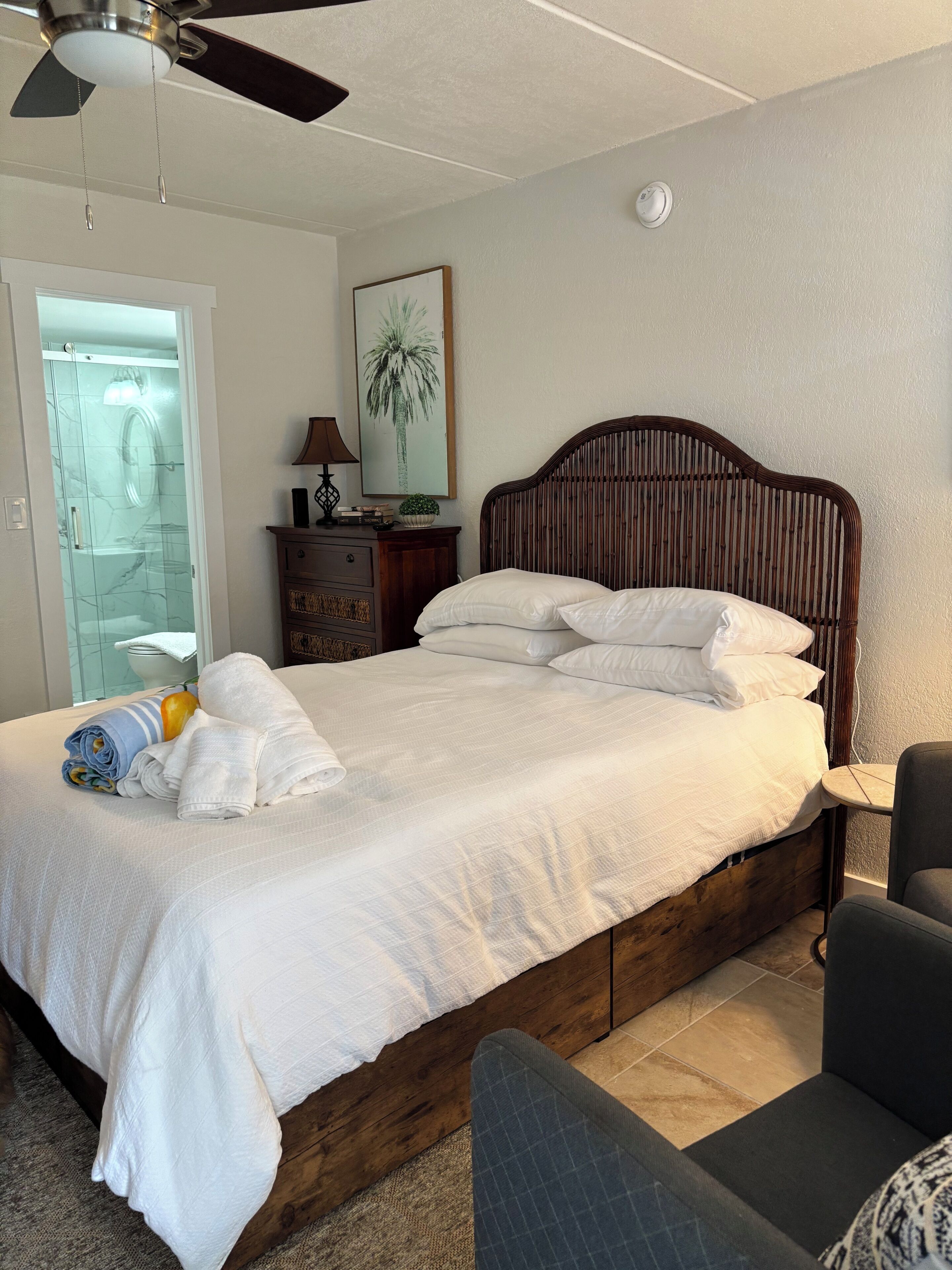 Un lit bien fait, avec du linge de lit et des serviettes blanches, une tête de lit en bois, une petite table de chevet avec une lampe et une chaise.