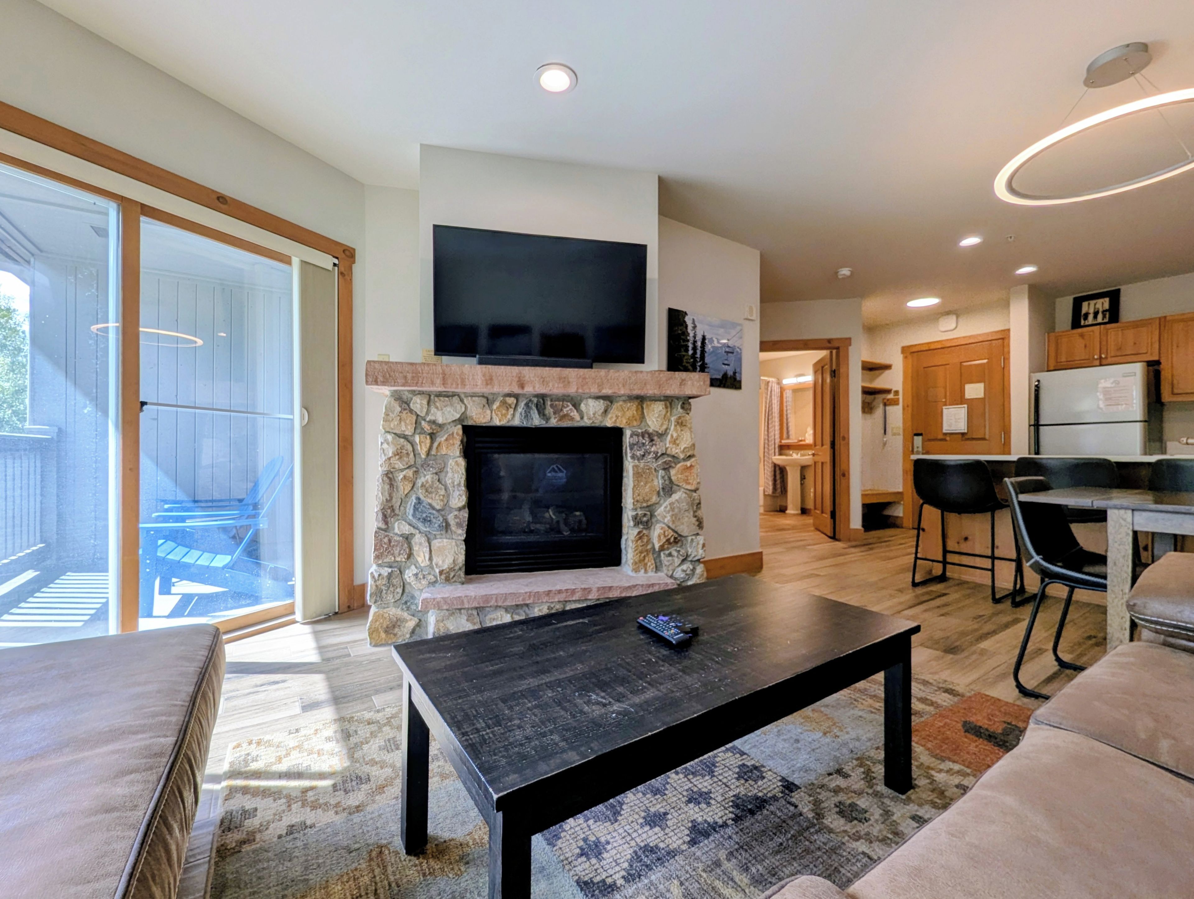 A modern living room with a stone fireplace, a flat-screen TV mounted above it, a dining area with bar stools, and a kitchen with wooden cabinets.