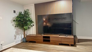 A modern living room with a large flat-screen TV mounted on a wooden entertainment unit, a potted plant, and a white door. - Lux 2BR Home Near Navy Base, AbbVie/Abbott, Six Flags, Beach (Waukegan)