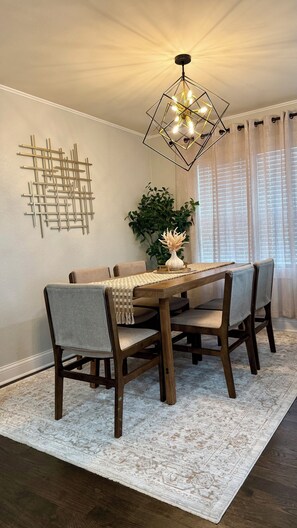 A dining area with a wooden table, chairs, a geometric wall art piece, a hanging light fixture, and a potted plant. - Lux 2BR Home Near Navy Base, AbbVie/Abbott, Six Flags, Beach (Waukegan)