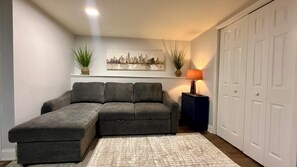 A grey sectional sofa with storage drawers, a patterned rug, a cityscape painting, a lamp, and a white door. - Lux 2BR Home Near Navy Base, AbbVie/Abbott, Six Flags, Beach (Waukegan)