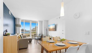 A modern living room with a sofa, a dining table set for four, and a large window offering a city view. - Cozy 1BR-Resort Lifestyle - Waterway View (Hallandale Beach)