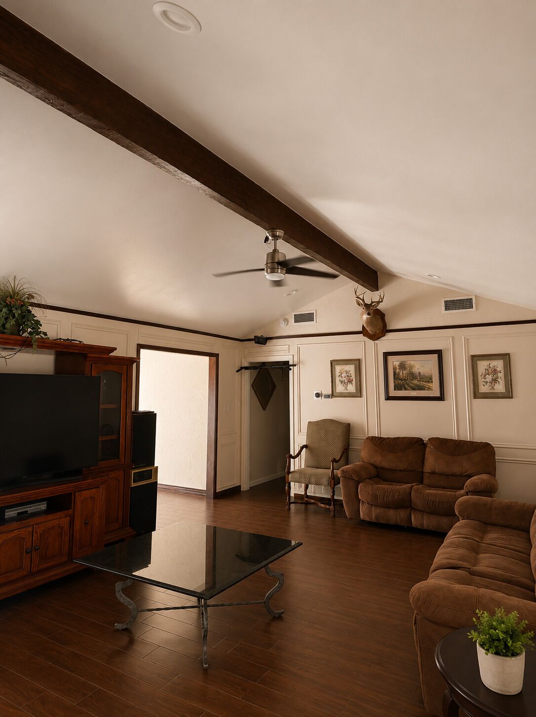 A spacious living room with a wooden floor, a glass coffee table, a brown sofa, a television, and a ceiling fan.
