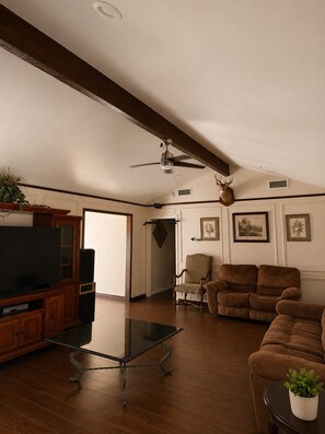 A spacious living room with a wooden floor, a glass coffee table, a brown sofa, a television, and a ceiling fan. - Spacious Getaway : 4BR-10Guest House Near AT&T Stadium, DFW Airport & Stockyards (Fort Worth)